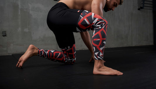 Athletic adult male in a low grappling stance on a dark training mat, wearing bold geometric-printed leggings