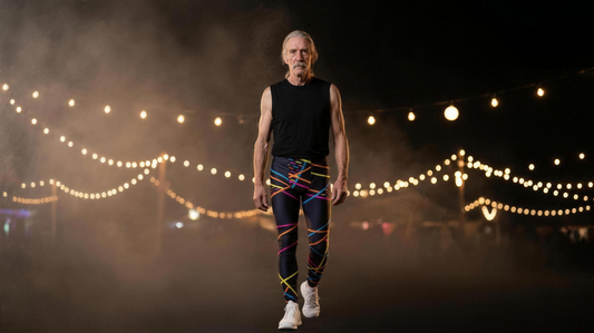 Man with long grey hair in black and rainbow geometric wrestling tights, walking forward under string lights at a night festival scene