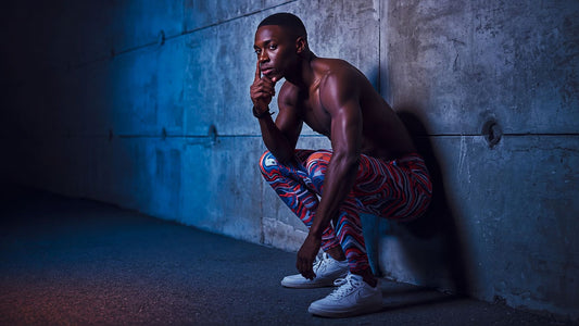 fashion editorial photograph of a male athlete in patterned leggings crouching near a concrete training wall, side-lit with subtle but moody directional neon lighting
