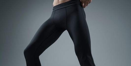 Wide horizontal editorial image showing the lower body of an adult man wearing men’s performance leggings, cropped from waist to mid-calf. Neutral studio background in soft grey tones, even lighting highlighting clean fit along thigh, knee, and calf.