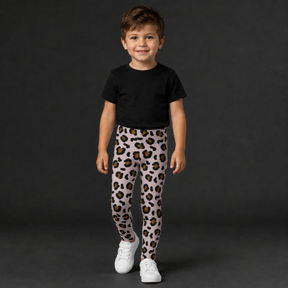 A young boy stands against a dark gray studio background wearing leopard-print leggings, a plain black t-shirt, and white sneakers.