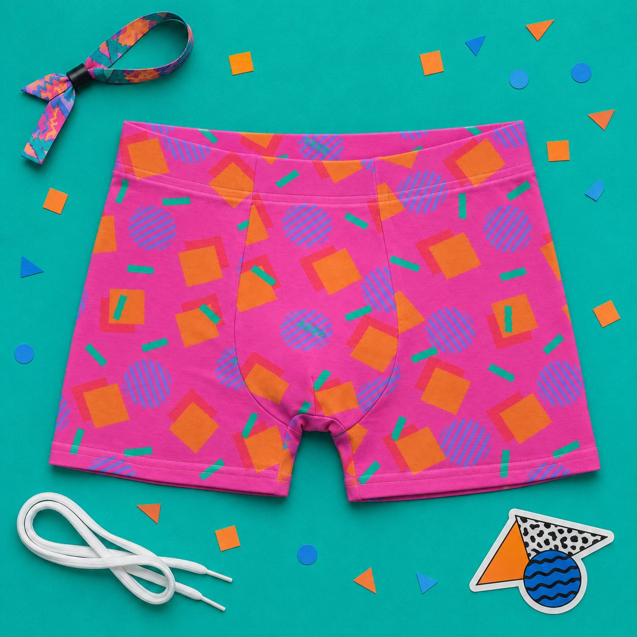 Pink boxer briefs with geometric pattern laid flat on a teal background with colorful 80s Memphis confetti and accessories.