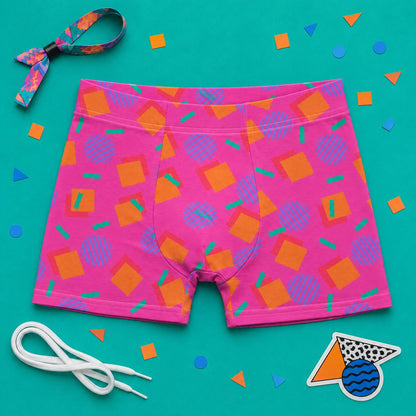Pink boxer briefs with geometric pattern laid flat on a teal background with colorful 80s Memphis confetti and accessories.