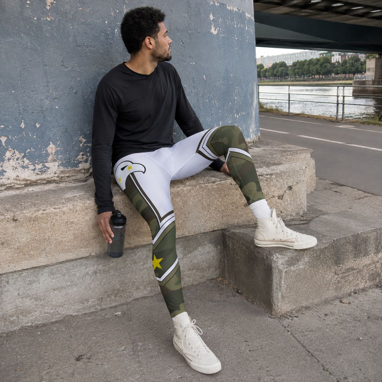 A man sits on concrete steps outdoors by a river, wearing patterned athletic leggings with a white upper leg, green camouflage lower leg, and a yellow star detail, paired with a black long-sleeve shirt and white sneakers.