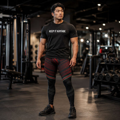 Man in a gym wearing a black t-shirt with 'KEEP IT KAYFABE' text and patterned leggings.