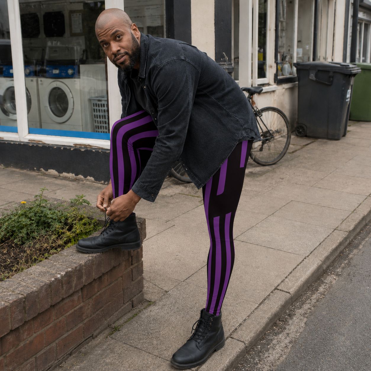A man wearing black and purple striped tights, a black denim jacket, and black boots bends down on a sidewalk to tie the laces of his right boot. A brick planter, a bicycle, and a storefront with washing machines are visible in the background.