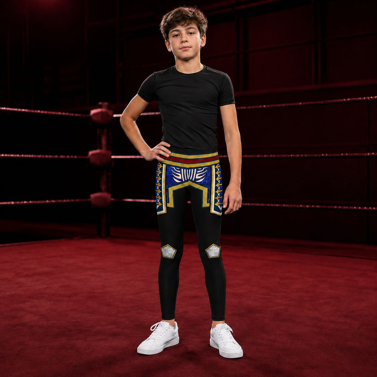 A boy stands in a boxing ring wearing a black short-sleeve athletic t-shirt and black cosplay wrestling tights in a retro American hero style with a colorful crocodile pattern on the upper thighs and blue knee patches, paired with white sneakers.