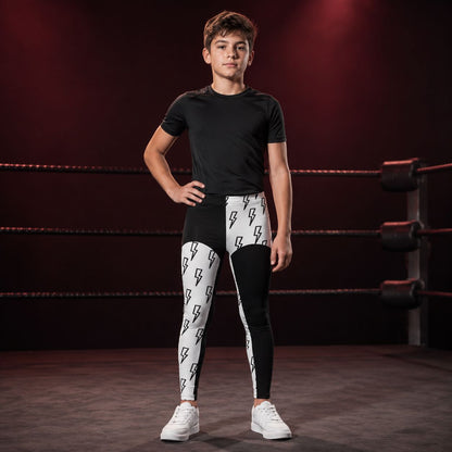 A young boy stands in a boxing ring wearing a black t-shirt and black-and-white lightning bolt cosplay wrestling tights with white sneakers.