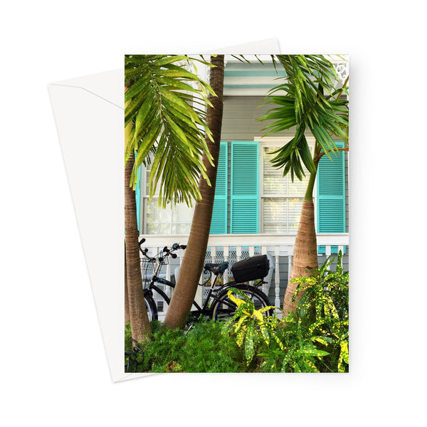 This greeting card shows a closeup of a traditional conch house with turquoise shutters beside some palms. A bicycle is parked next to the white fence. There are plants in the foreground. 