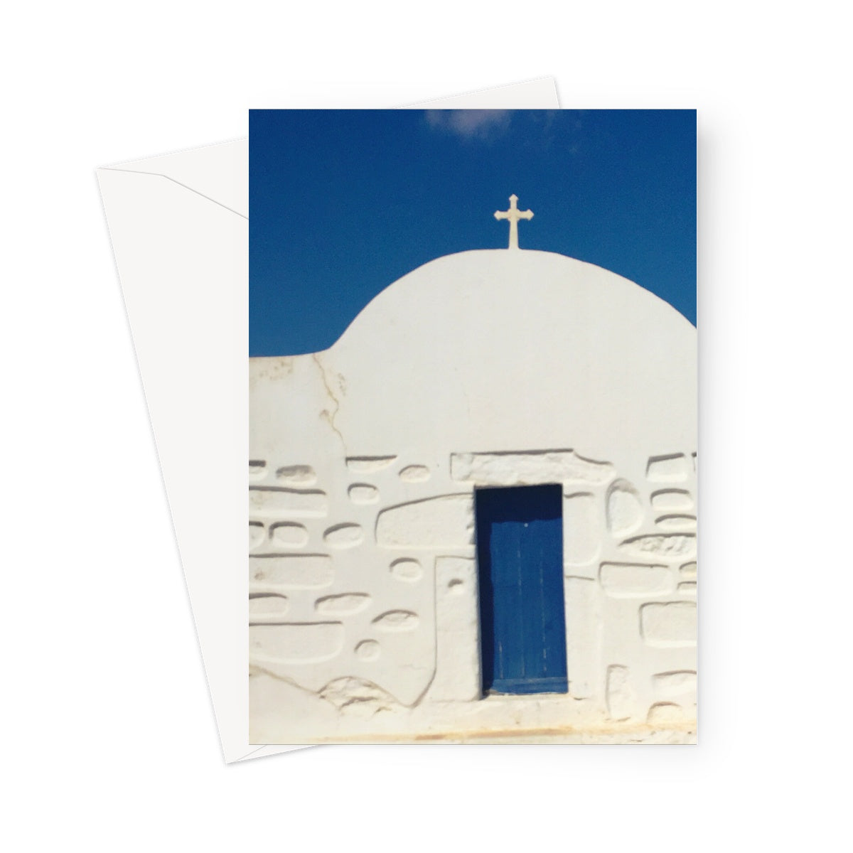 Greeting card showing a small whitewashed traditional Greek Orthodox church. The walls are carved in the traditional detail of the Greek islands. A blue entrance door matches the blue sky.