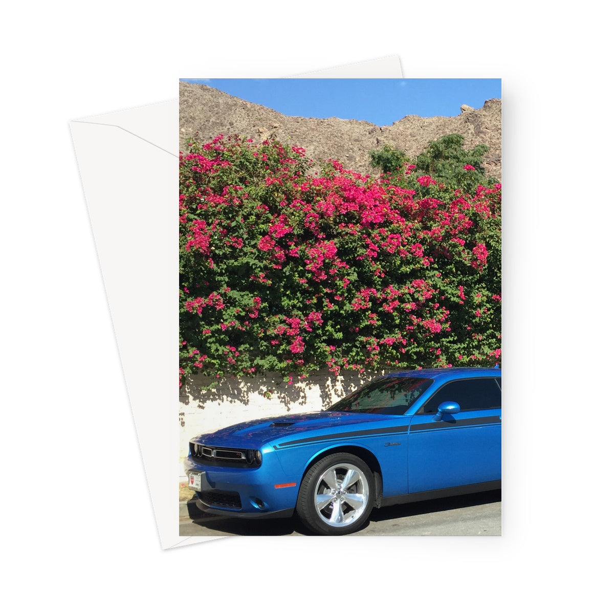 Blue Mustang car parked against a pale wall, which is overhung with a cerise flowering bush. In the distance are the brown mountains and azure blue sky of Palm Springs, California, in this greeting card
