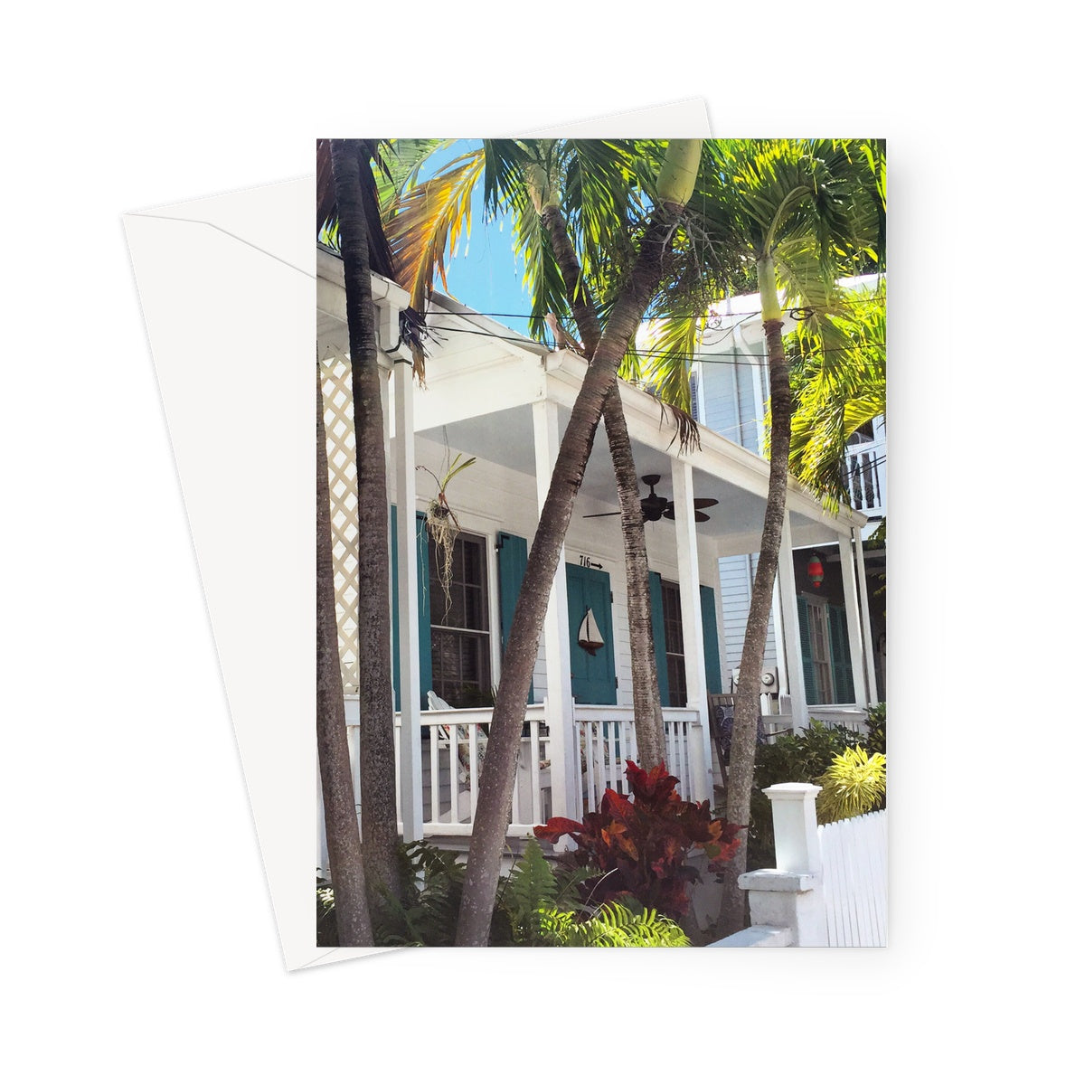 This greeting card shows the white veranda of a conch house with green/blue shutters and front door. There are palms immediately outside in the front garden. A small white yacht is attached to the front door.