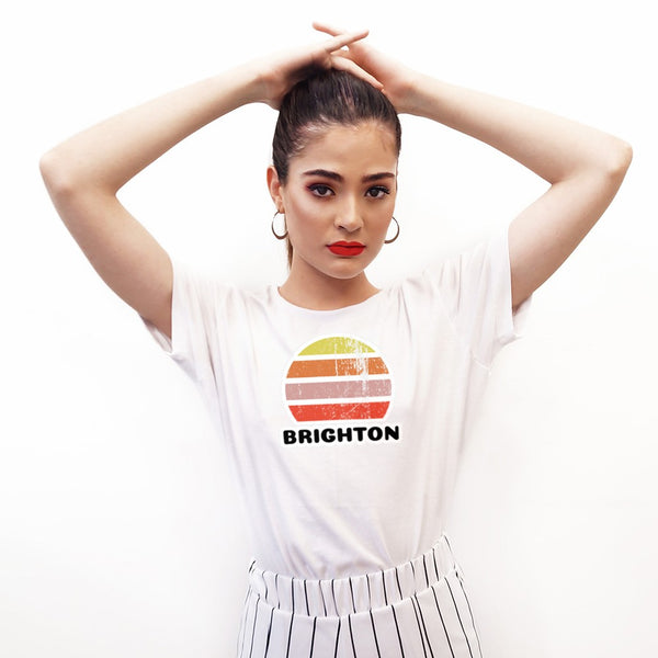 Features a distressed abstract retro sunset graphic in yellow, orange, pink and scarlet stripes rising up from the famous Brighton place name in this white cotton  t-shirt