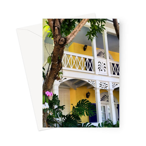 This greeting card shows a deep yellow painted traditional conch house with detailed white veranda and shutters. There is a pink orchid and Monstera plant in the front garden