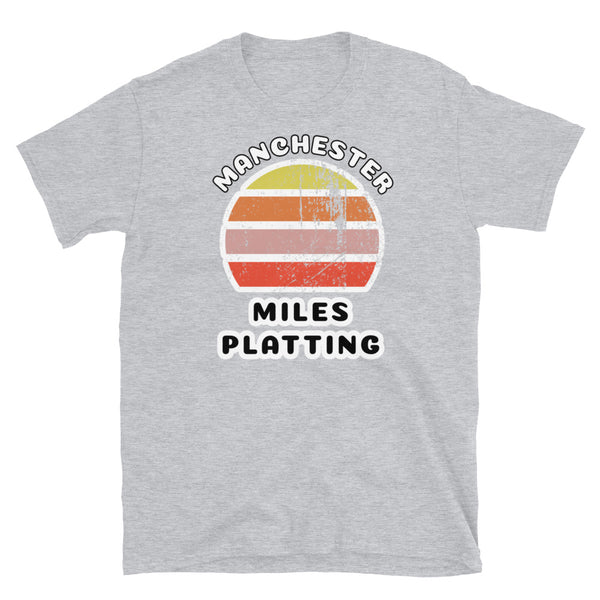 Distressed style abstract retro sunset graphic in yellow, orange, pink and scarlet stripes. The name of Manchester is displayed at the top wrapped around the sunset. Below the retro sunset design is the famous Manchester place name of Miles Platting on this light grey cotton t-shirt.